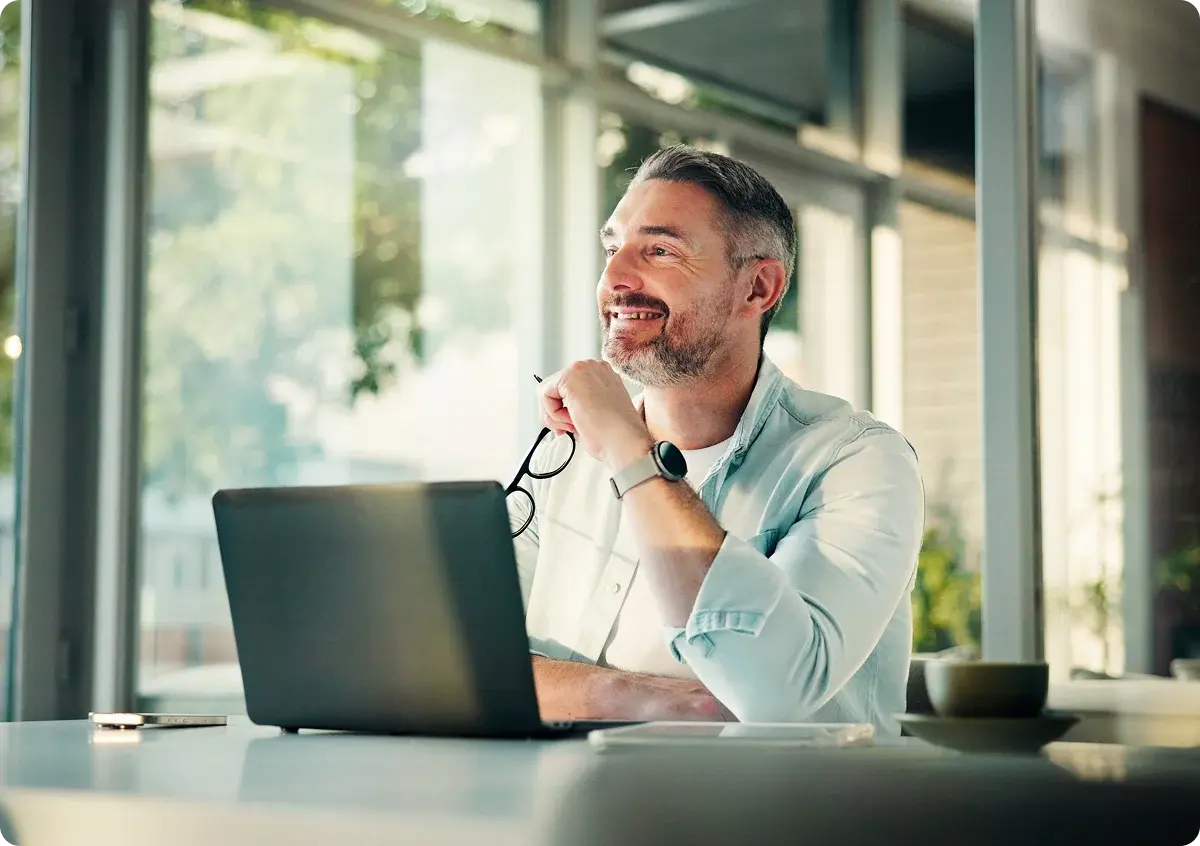 man-with-glasses-in-hand-sitting-at-desk-looking-out-window-and-smiling man-with-glasses-in-hand-sitting-at-desk-looking-out-window-and-smiling